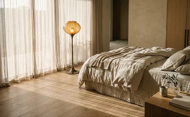 A bedroom in low sun, a glass lamp glowing beside the bed.